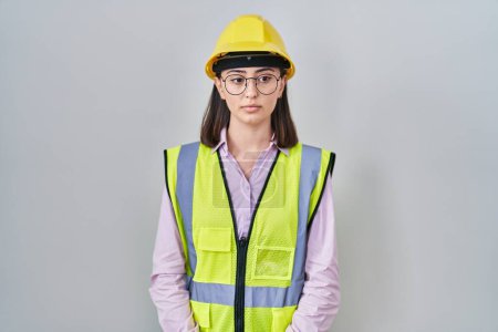 Photo for Hispanic girl wearing builder uniform and hardhat relaxed with serious expression on face. simple and natural looking at the camera. - Royalty Free Image
