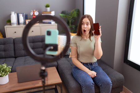 Photo for Beautiful brunette woman recording vlog tutorial with smartphone at home scared and amazed with open mouth for surprise, disbelief face - Royalty Free Image
