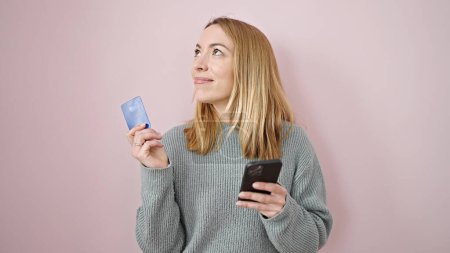Foto de Mujer rubia joven de compras con teléfono inteligente y tarjeta de crédito pensando en el fondo rosa aislado - Imagen libre de derechos
