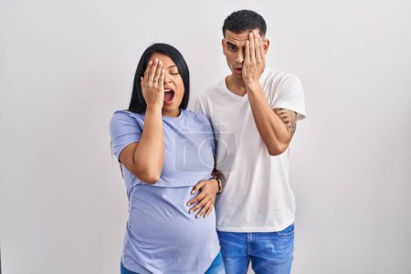 Photo for Young hispanic couple expecting a baby standing over background yawning tired covering half face, eye and mouth with hand. face hurts in pain. - Royalty Free Image