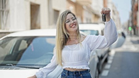 Téléchargez les photos : Jeune femme blonde souriante confiante tenant la clé de la nouvelle voiture à la rue - en image libre de droit