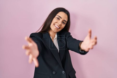 Téléchargez les photos : Jeune femme brune portant un style d'affaires sur fond rose regardant la caméra souriant à bras ouverts pour un câlin. expression joyeuse embrassant le bonheur. - en image libre de droit
