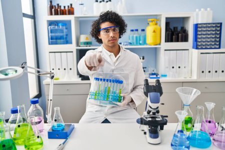 Photo for Hispanic man with curly hair working at scientist laboratory pointing with finger to the camera and to you, confident gesture looking serious - Royalty Free Image