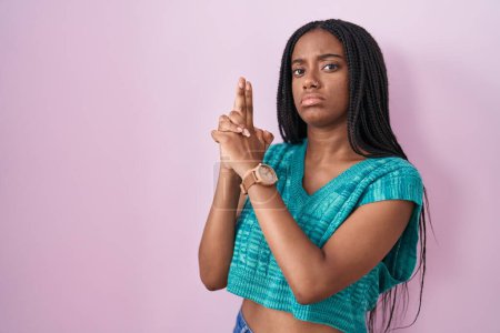Téléchargez les photos : Jeune afro-américain avec des tresses debout sur fond rose tenant un pistolet symbolique avec geste de la main, jouant tuer des armes de tir, visage en colère - en image libre de droit