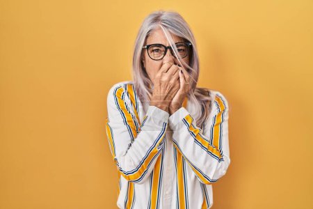 Téléchargez les photos : Femme d'âge moyen avec les cheveux gris debout sur fond jaune portant des lunettes sentant quelque chose de puant et dégoûtant, odeur intolérable, retenant la respiration avec les doigts sur le nez. mauvaise odeur - en image libre de droit