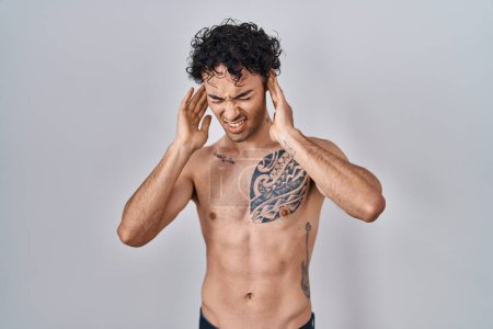 Foto de Hombre hispano de pie sin camisa cubriendo oídos con dedos con expresión molesta por el ruido de la música alta. concepto de sordo. - Imagen libre de derechos