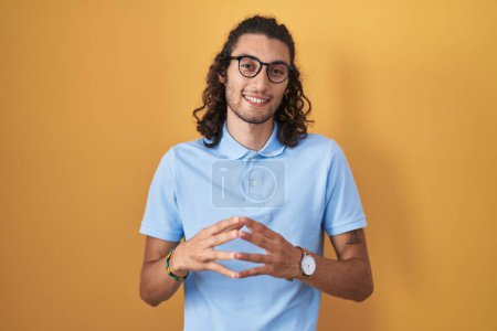 Téléchargez les photos : Jeune homme hispanique debout sur fond jaune mains ensemble et doigts croisés souriant détendu et joyeux. succès et optimisme - en image libre de droit