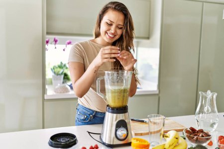 Photo for Young beautiful hispanic woman preparing vegetable smoothie with blender at the kitchen - Royalty Free Image