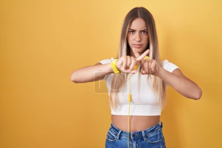 Foto de Mujer rubia joven de pie sobre fondo amarillo con auriculares expresión de rechazo cruzando los dedos haciendo signo negativo - Imagen libre de derechos