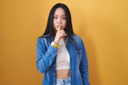 Téléchargez les photos : Jeune femme asiatique debout sur fond jaune demandant à être calme avec le doigt sur les lèvres. silence et concept secret. - en image libre de droit