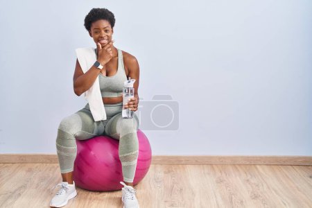 Photo for African american woman wearing sportswear sitting on pilates ball looking confident at the camera smiling with crossed arms and hand raised on chin. thinking positive. - Royalty Free Image