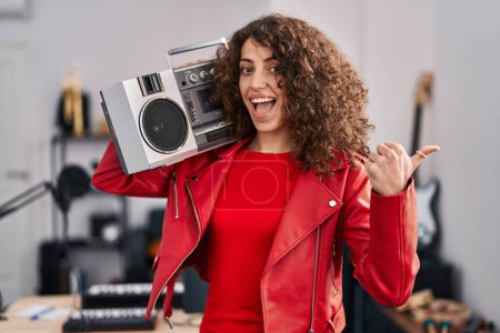 Foto de Mujer hispana con el pelo rizado sosteniendo boombox, escuchando música apuntando con el pulgar hacia el lado sonriendo feliz con la boca abierta - Imagen libre de derechos
