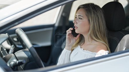 Photo for Young blonde woman talking on smartphone sitting on car at street - Royalty Free Image
