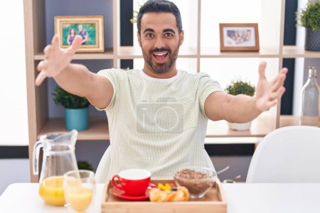 Téléchargez les photos : Homme hispanique avec barbe petit déjeuner en regardant la caméra souriant à bras ouverts pour un câlin. expression joyeuse embrassant le bonheur. - en image libre de droit
