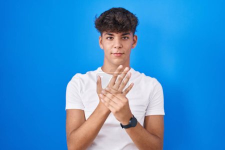 Téléchargez les photos : Adolescent hispanique debout sur fond bleu souffrant de douleur aux mains et aux doigts, inflammation de l'arthrite - en image libre de droit