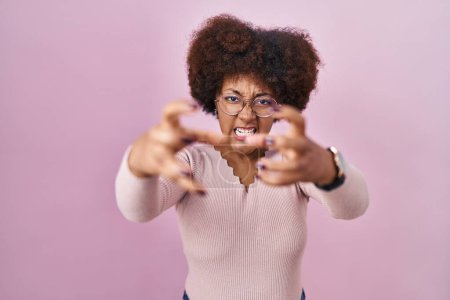 Foto de Joven mujer afroamericana de pie sobre fondo rosa gritando frustrado con rabia, las manos tratando de estrangular, gritando loco - Imagen libre de derechos