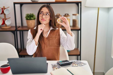 Téléchargez les photos : Jeune femme brune travaillant avec un ordinateur portable tenant monnaie virtuelle Bitcoin visage sérieux penser à la question avec la main sur le menton, réfléchi sur l'idée déroutante - en image libre de droit