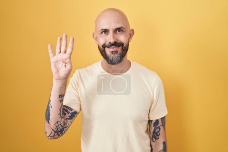 Foto de Hombre hispano con tatuajes de pie sobre fondo amarillo mostrando y apuntando hacia arriba con los dedos número cuatro mientras sonríe confiado y feliz. - Imagen libre de derechos