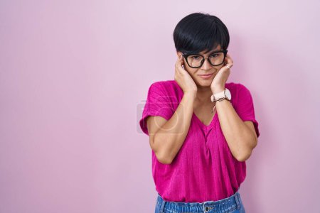 Foto de Mujer asiática joven con el pelo corto de pie sobre el fondo rosa que cubre las orejas con los dedos con expresión molesta por el ruido de la música fuerte. concepto de sordo. - Imagen libre de derechos