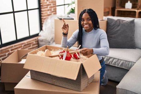 Téléchargez les photos : Femme afro-américaine se déplaçant vers une nouvelle maison boîtes d'ouverture souriant heureux pointant avec la main et le doigt sur le côté - en image libre de droit