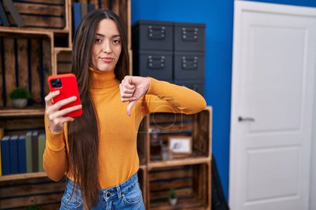 Téléchargez les photos : Jeune femme brune utilisant un smartphone avec un visage en colère, signe négatif montrant aversion avec les pouces vers le bas, concept de rejet - en image libre de droit