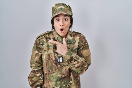 Téléchargez les photos : Jeune femme sud-asiatique portant uniforme camouflage armée surpris pointant du doigt sur le côté, bouche ouverte expression étonnée. - en image libre de droit