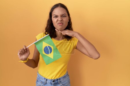Téléchargez les photos : Jeune femme hispanique tenant drapeau brésilien tranchant la gorge avec la main comme couteau, menace d'agression avec une violence furieuse - en image libre de droit