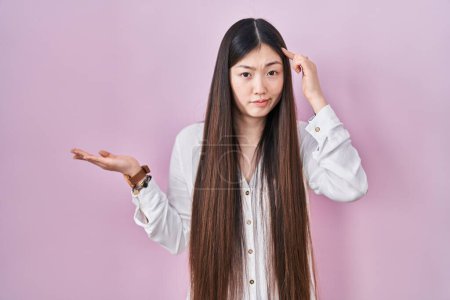 Photo for Chinese young woman standing over pink background confused and annoyed with open palm showing copy space and pointing finger to forehead. think about it. - Royalty Free Image