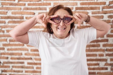 Téléchargez les photos : Femme âgée avec des lunettes debout sur le mur de briques faisant symbole de paix avec les doigts sur le visage, souriant joyeuse montrant la victoire - en image libre de droit
