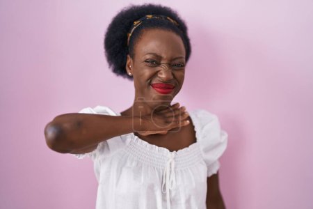 Foto de Mujer africana con el pelo rizado de pie sobre el fondo rosa cortando la garganta con la mano como cuchillo, amenazan la agresión con violencia furiosa - Imagen libre de derechos