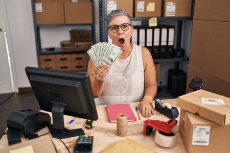 Photo for Middle age woman working at small business ecommerce holding dollars scared and amazed with open mouth for surprise, disbelief face - Royalty Free Image