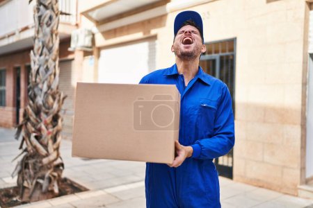 Photo for Young hispanic man delivering box angry and mad screaming frustrated and furious, shouting with anger looking up. - Royalty Free Image