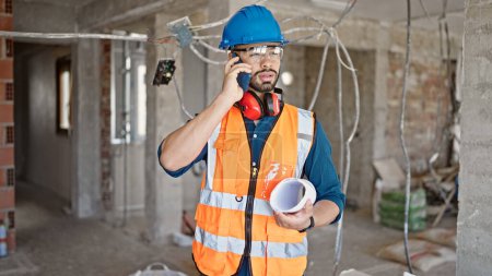 Photo for Young hispanic man builder standing with blueprints speaking on the phone at construction site - Royalty Free Image