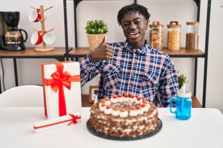 Téléchargez les photos : Homme africain avec dreadlocks célébrant anniversaire tenant grand gâteau au chocolat souriant heureux et positif, pouce levé faire excellent et signe d'approbation - en image libre de droit