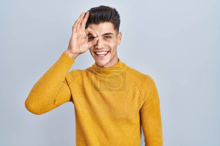 Téléchargez les photos : Jeune homme hispanique debout sur fond bleu faisant un geste correct avec la main souriante, les yeux regardant à travers les doigts avec un visage heureux. - en image libre de droit