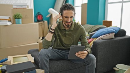 Foto de Hombre hispano joven hablando en smartphone usando touchpad en nuevo hogar - Imagen libre de derechos