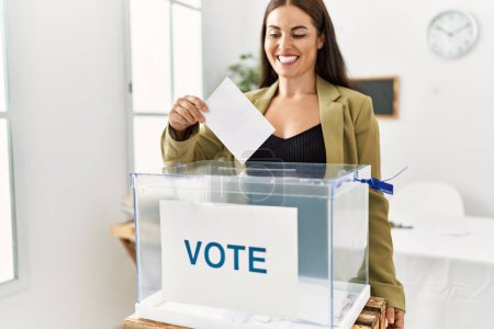Photo for Young beautiful hispanic woman smiling confident putting vote on ballot box at electoral college - Royalty Free Image