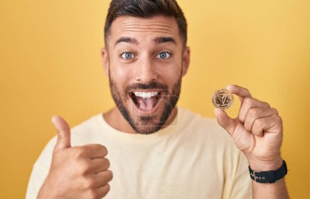 Foto de Handsome hispanic man holding tron cryptocurrency coin smiling happy and positive, thumb up doing excellent and approval sign - Imagen libre de derechos