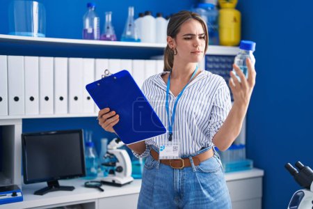Téléchargez les photos : Jeune femme scientifique lisant un document tenant une bouteille au laboratoire - en image libre de droit