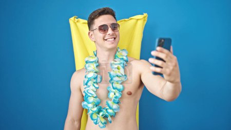 Foto de Joven turista hispano hace selfie por teléfono inteligente relajado en hamaca inflable de la piscina sobre fondo azul aislado - Imagen libre de derechos