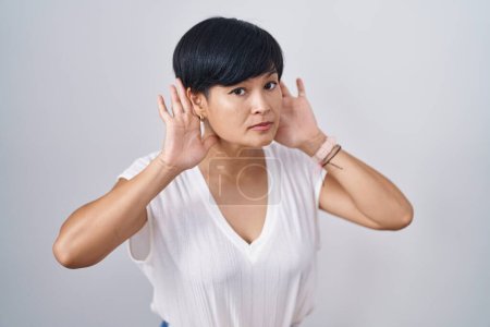 Téléchargez les photos : Jeune femme asiatique aux cheveux courts debout sur fond isolé essayant d'entendre les deux mains sur le geste de l'oreille, curieuse de commérages. problème auditif, sourd - en image libre de droit
