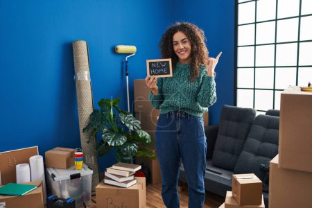 Foto de Joven mujer hispana sosteniendo pizarra con nuevo texto en casa sonriendo feliz señalando con la mano y el dedo hacia un lado - Imagen libre de derechos