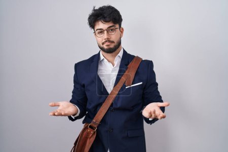 Foto de Hombre hispano con barba vistiendo ropa de negocios despistado y confundido con los brazos abiertos, sin concepto de idea. - Imagen libre de derechos