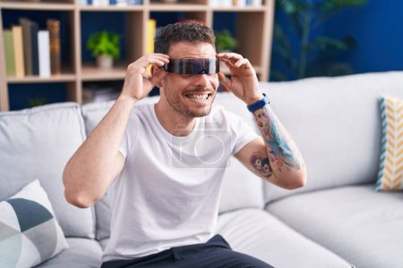 Téléchargez les photos : Jeune homme hispanique jouant à un jeu vidéo en utilisant des lunettes de réalité virtuelle à la maison - en image libre de droit
