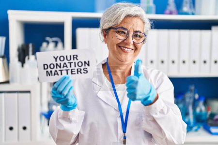 Téléchargez les photos : Femme d'âge moyen avec les cheveux gris travaillant au laboratoire scientifique tenant vos questions de don bannière souriant heureux et positif, pouce levé faire excellent et signe d'approbation - en image libre de droit