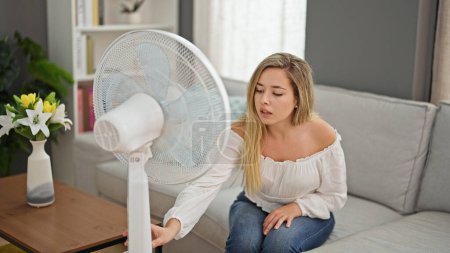 Photo for Young blonde woman sitting on sofa suffering for hot starting fan at home - Royalty Free Image