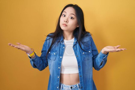 Foto de Mujer asiática joven de pie sobre fondo amarillo expresión despistada y confusa con los brazos y las manos levantadas. concepto de duda. - Imagen libre de derechos
