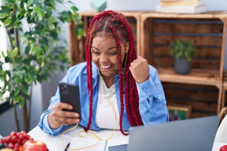Téléchargez les photos : Femme afro-américaine avec les cheveux tressés en utilisant smartphone assis sur la table criant fier, célébrant la victoire et le succès très excité avec le bras levé - en image libre de droit