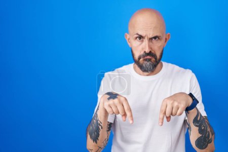 Téléchargez les photos : Homme hispanique avec des tatouages debout sur fond bleu pointant vers le bas regardant triste et contrarié, indiquant la direction avec les doigts, malheureux et déprimé. - en image libre de droit