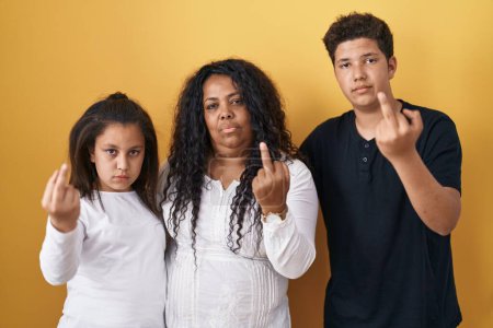 Foto de Family of mother, daughter and son standing over yellow background showing middle finger, impolite and rude fuck off expression - Imagen libre de derechos